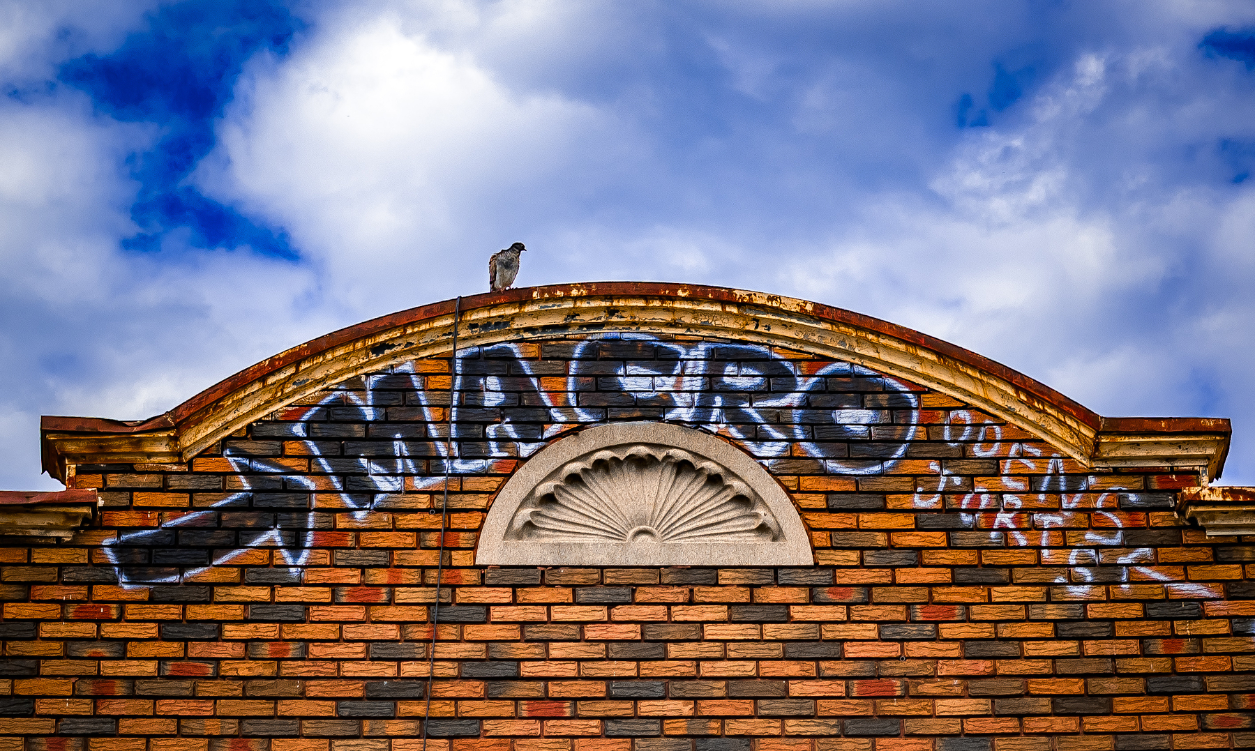 Auto-generated description: A brick building facade features graffiti with the words MACRO OPEN 7 DAYS against a partly cloudy sky.